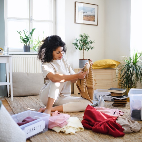 A young woman sitting on the floor, sorting clothes from her wardrobe at home, carefully preparing items for donation.