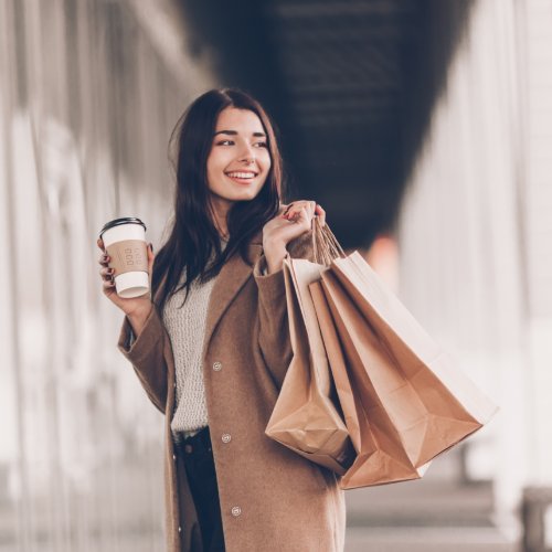 A woman wearing sunglasses, carrying shopping bags and a coffee cup, while walking outdoors near storefront windows.
