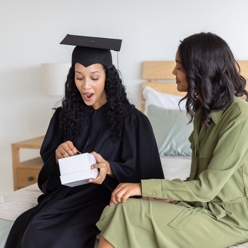 A graduate in a cap and gown holds an open gift box as she sits by a loved one. Her mouth is open in surprise.