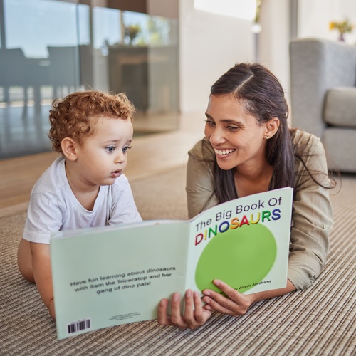 A woman and a young child sitting on a living room floor while looking at an open dinosaur book together.