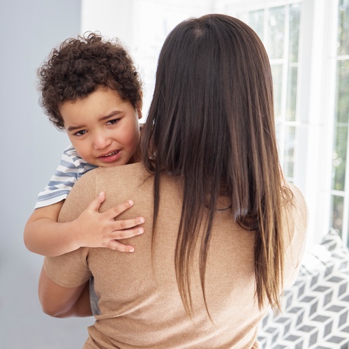 The backside of a woman standing and holding a crying child against her shoulder inside a bright living room.