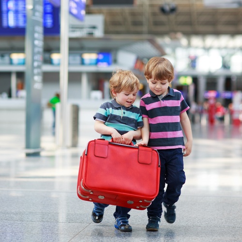Two young children in striped shirts hold a large red suitcase together while walking in an airport terminal.