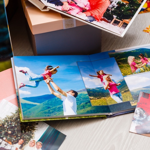 An open photo album rests on a wooden surface beside scattered photographs, a storage box, and other crafting materials.
