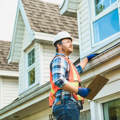 A man wearing a hard hat and safety vest inspects the roof of a home. He wears blue gloves and holds a clipboard.