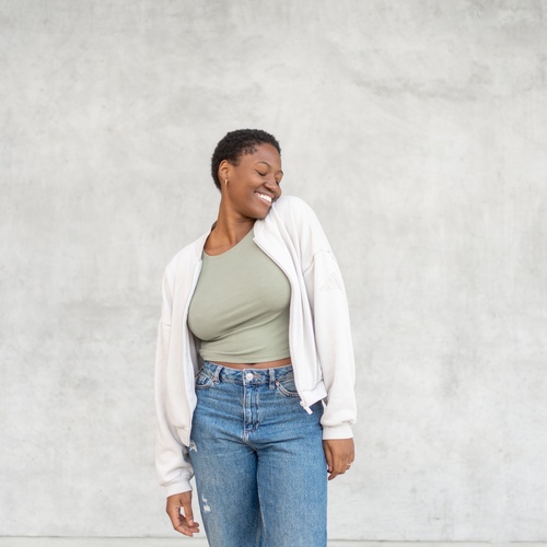 Smiling woman wearing a casual cardigan, crop top, and jeans while posing against a plain gray wall.