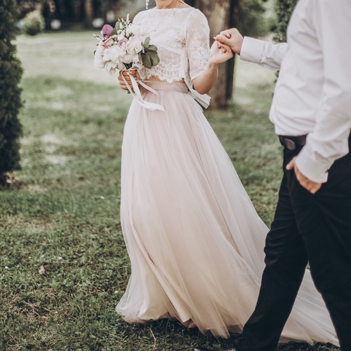 A woman in a lace top and long tulle skirt holding a bouquet while walking hand in hand with a man outside.