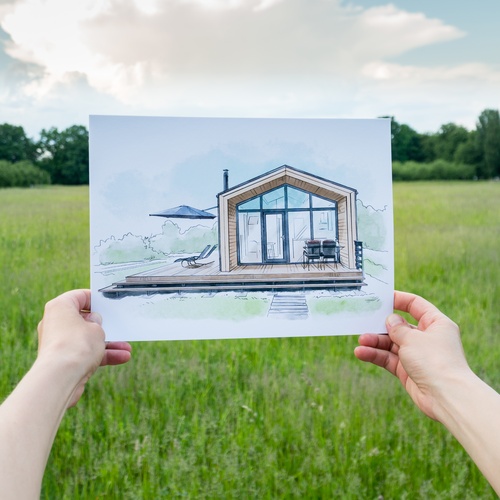 Someone holds a painted sketch of a home in front of a chunk of empty land with tall grass growing.