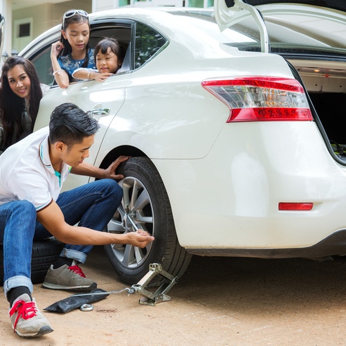 A man uses a socket wrench to remove a tire screw from a vehicle. A woman and two kids watch from inside the car.