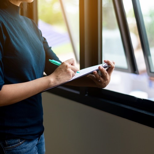 A close-up of a woman writing notes on a clipboard while standing beside an open window during a property inspection.