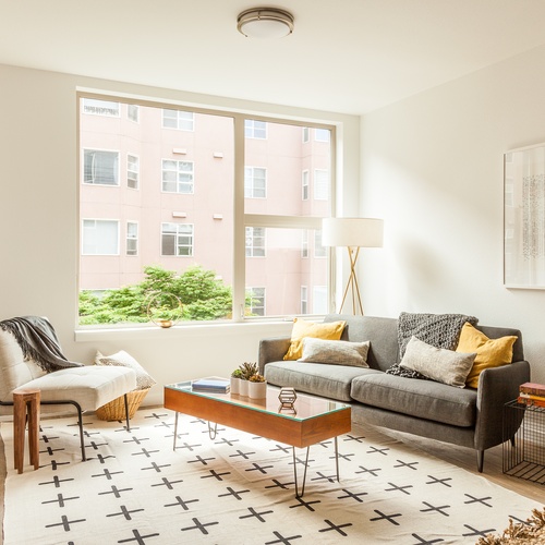 A living room features a gray couch, a white chair, a coffee table, a rug, a large window, a lamp, and metal crates.