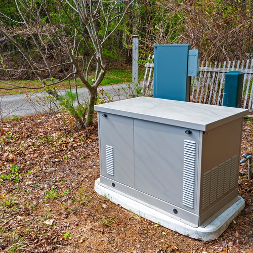 A close-up of a residential standby generator in the front yard of a home next to a fence and a driveway.