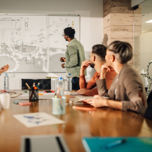 A group of workers sits at a meeting table. One of them stands at a whiteboard with ideas written on it.
