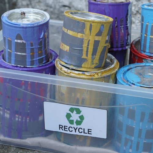 Paint-splattered cans sit inside a clear plastic recycling bin labeled "RECYCLE" with a green recycling symbol.