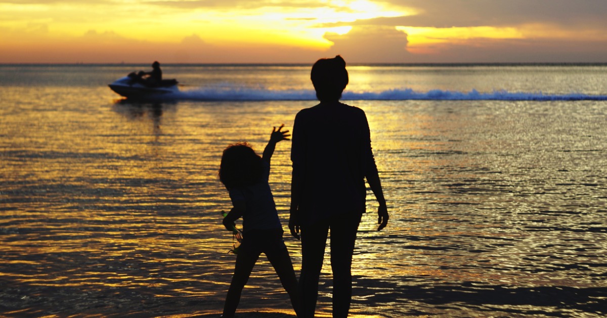 Two silhouetted people standing in shallow water at sunset as a personal watercraft passes in the distance.