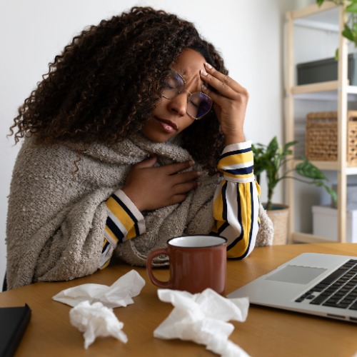 A close-up of a woman sitting at a desk with a laptop and tissues while resting one hand on her forehead indoors.