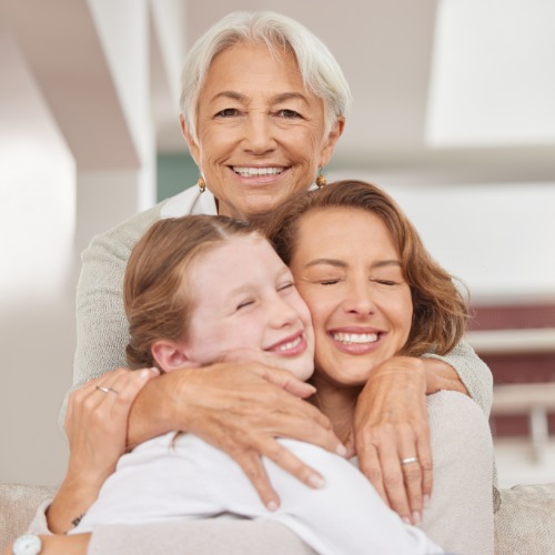 A grandmother, a mother, and a young girl are wearing neutral clothing while smiling and hugging tightly.