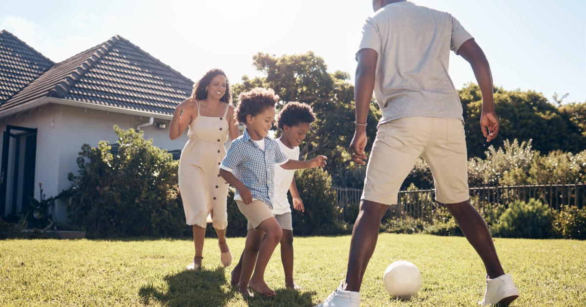 A family playing soccer together on a sunny backyard lawn, with two children chasing the ball near their parents.