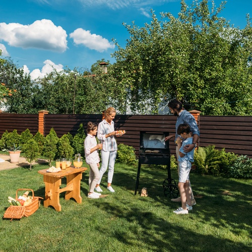 A family of four gathered around an outdoor grill in a backyard with a pool, picnic basket, and food table.