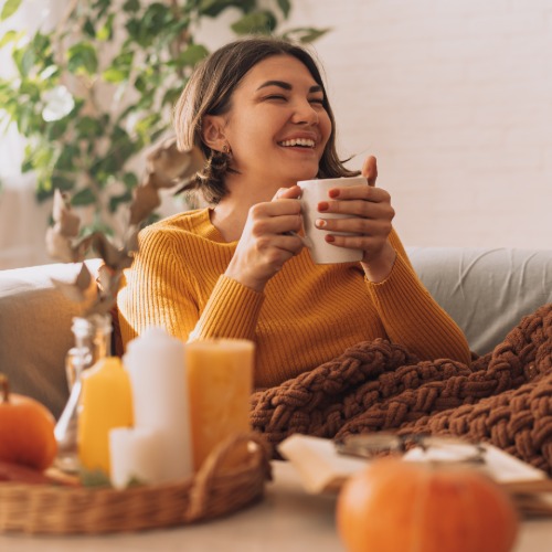 A young woman sits on the couch at home as she smiles and laughs, holding a hot mug with steam coming out of it.