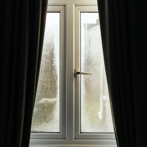 A white double-pane window with condensation on both panes, dark curtains on either side, and a central handle.