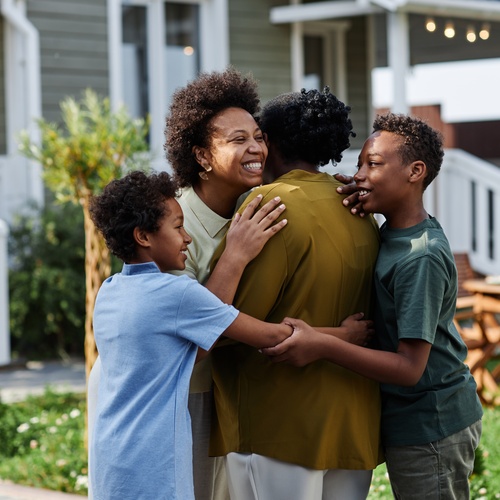 A smiling woman and two children hugging a person in a yard, with adults standing blurred near a house.