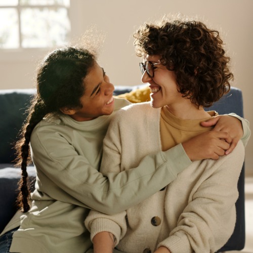Mom and daughter sit closely on a sofa, smiling at each other as one wraps an arm around the other's shoulders.