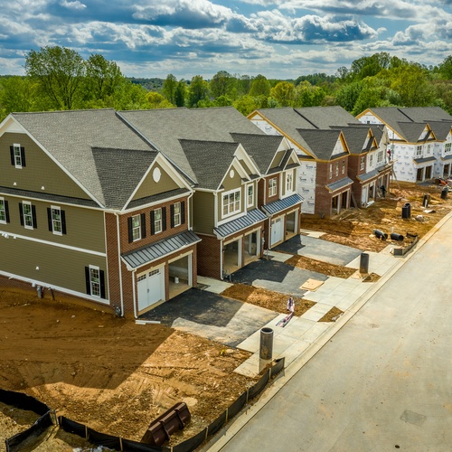 A residential street features new townhomes that are at different stages of construction. A forest is behind the homes.