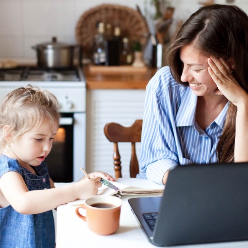 A woman sits at a kitchen table with an open laptop and a cup of coffee. A young child draws in a notebook at the table.