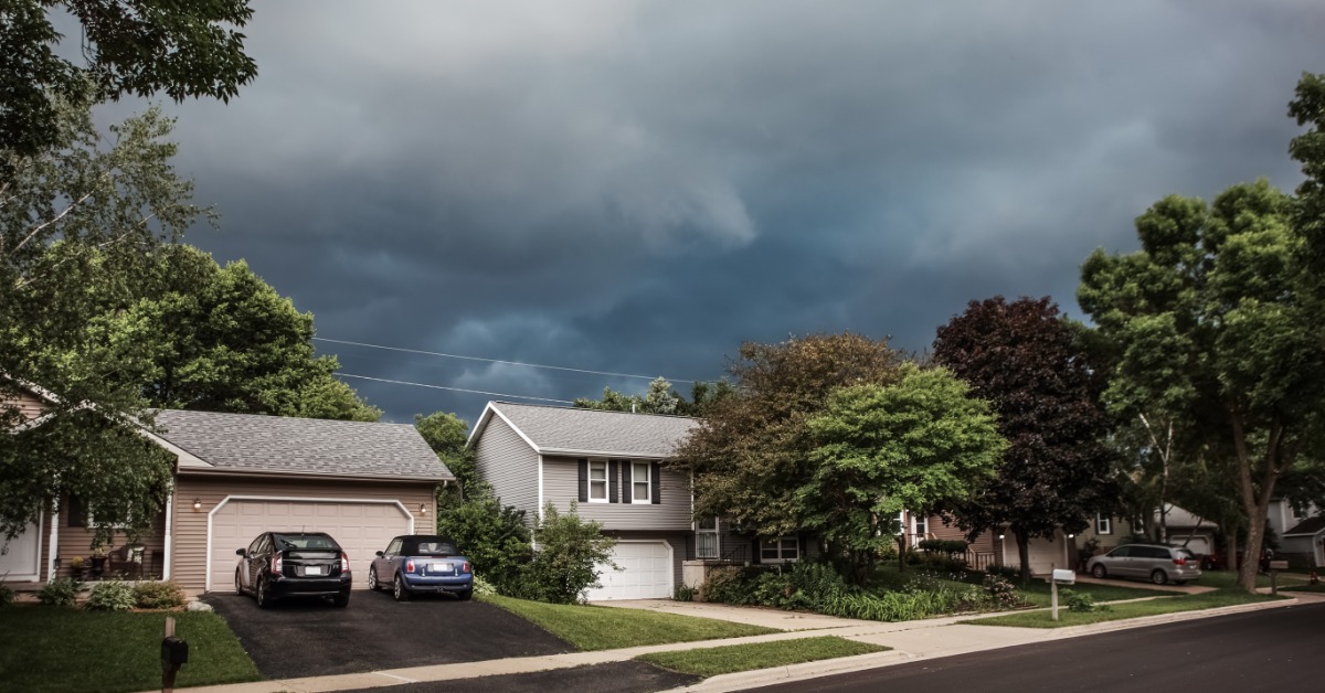 Dark gray storm clouds swirling above a suburban neighborhood during the daytime. The neighborhood has many large trees.