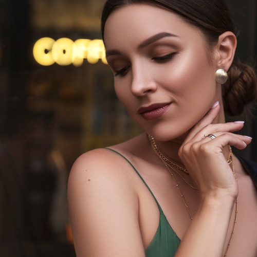 A dark-haired woman with closed eyes touching her jaw, wearing gold earrings and a green top with a black jacket.