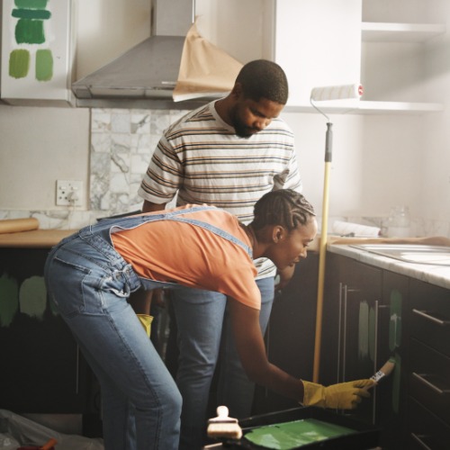 A couple stands in their kitchen as they complete cabinetry updates with paint. There are green swatches on the wall.