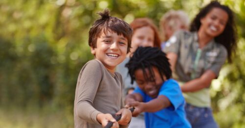 Smiling children play tug-of-war outdoors, with a boy in the foreground pulling a rope as others join behind him.