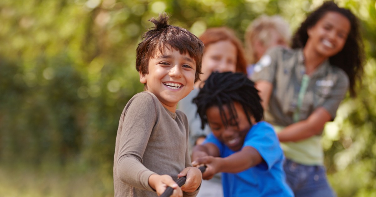 Smiling children play tug-of-war outdoors, with a boy in the foreground pulling a rope as others join behind him.