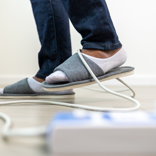 A person wearing gray slippers in their living room tripping over a long, white wire attached to a power strip.